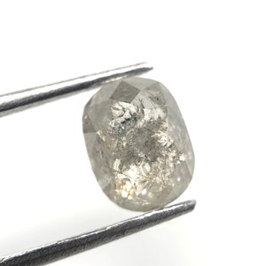 May include: A faceted, oval-shaped, gray diamond held by silver tweezers. The diamond has internal inclusions and a slightly translucent appearance, with a mix of light and dark gray tones. The background is white.