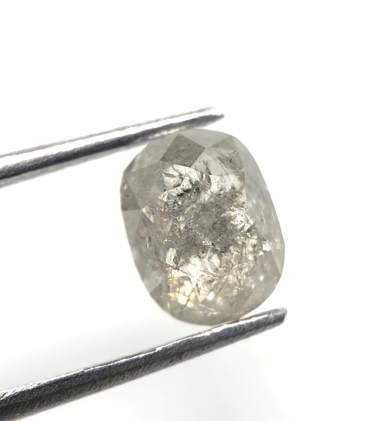 May include: A faceted, oval-shaped, gray diamond held by silver tweezers. The diamond has internal inclusions and a slightly translucent appearance, with a mix of light and dark gray tones. The background is white.