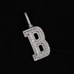 Könnte beinhalten: Ein goldfarbener Anhänger in Form des Buchstabens "B", besetzt mit zahlreichen kleinen, klaren Edelsteinen. Der Anhänger hat eine Schlaufe für eine Kette und ist vor schwarzem Hintergrund platziert.