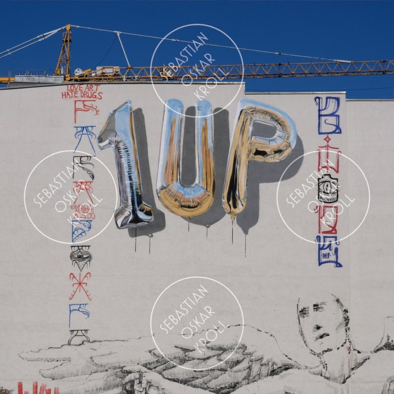 K&ouml;nnte beinhalten: Ein Wandgem&auml;lde vor blauem Himmel zeigt silberne und goldene Folienballons mit der Aufschrift "1UP". Das Kunstwerk enth&auml;lt den Text "LOVE ART HATE DRUGS" und stilisierte vertikale Schrift in Rot, Blau und Schwarz. Im Hintergrund ist ein Baukran zu sehen.