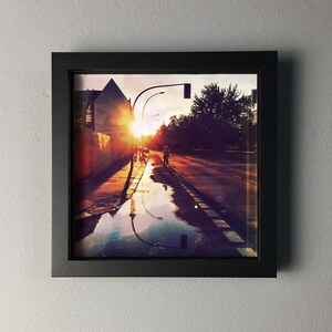May include: Framed photograph of a city street at sunset. The sun's rays burst through the buildings, reflecting in puddles on the wet asphalt. A person is riding a bicycle down the street. The frame is black.