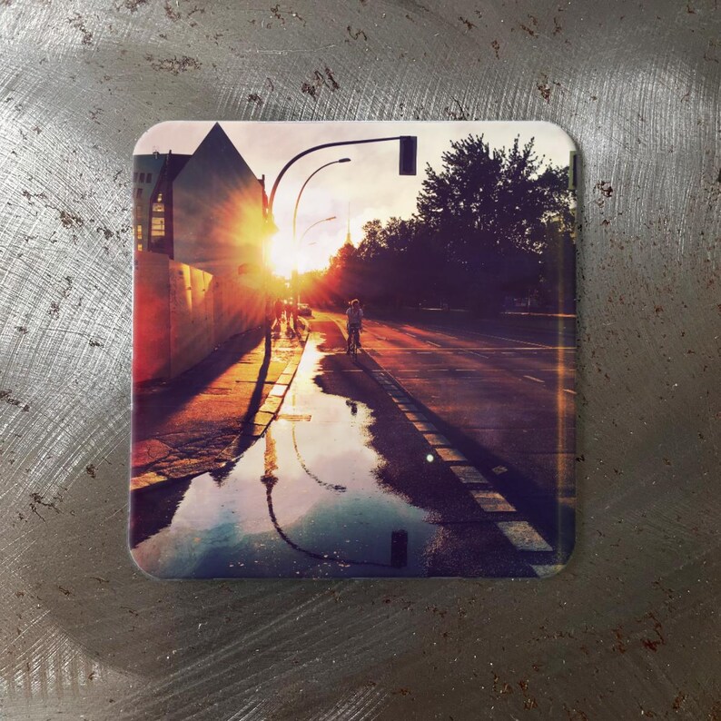 May include: Square coaster featuring a vibrant sunset scene over a city street. The image shows a person on a bicycle, a building, and a reflection in a puddle. The color palette includes warm oranges, yellows, and blues.