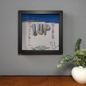 K&ouml;nnte beinhalten: Gerahmtes Kunstwerk mit dem Wort "1UP" in silbernen Ballonbuchstaben auf einer wei&szlig;en Wand mit Graffiti und einem Baukran im Hintergrund. Eine Skizze einer Person befindet sich unten. Der Rahmen ist schwarz.