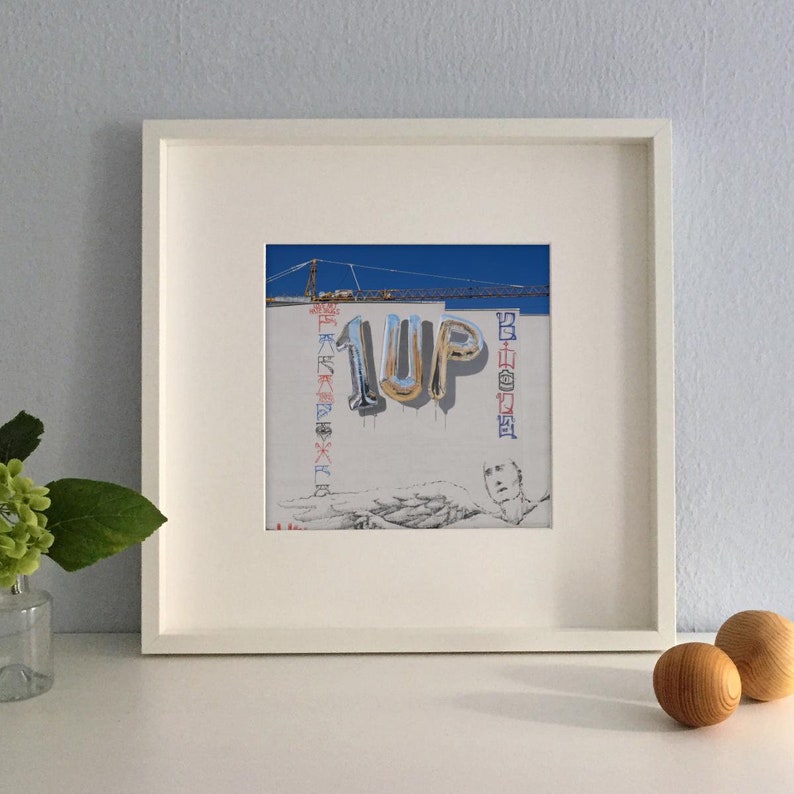 K&ouml;nnte beinhalten: Gerahmter Kunstdruck mit einem Graffiti-Mural mit dem Text "1UP" in silbernen Ballonbuchstaben. Das Kunstwerk zeigt einen Kran, ein Portr&auml;t und andere farbenfrohe grafische Elemente vor einer wei&szlig;en Wand, gerahmt in einem wei&szlig;en Quadrat.