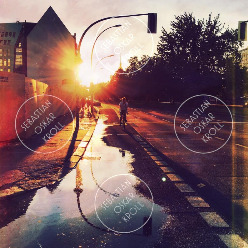 May include: A street scene at sunset with a cyclist on a road reflecting the sky and buildings. The image has a warm, golden hue from the setting sun, with the text "SEBASTIAN OSKAR KROLL" in a circle overlaying the image.