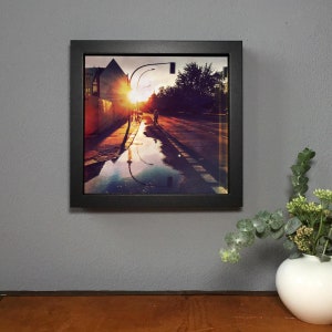 May include: A framed photograph captures a sunset over a wet street, with the sun's rays bursting through buildings. The image reflects in a large puddle, with a person walking in the distance. The frame is black, and the overall mood is warm and inviting.