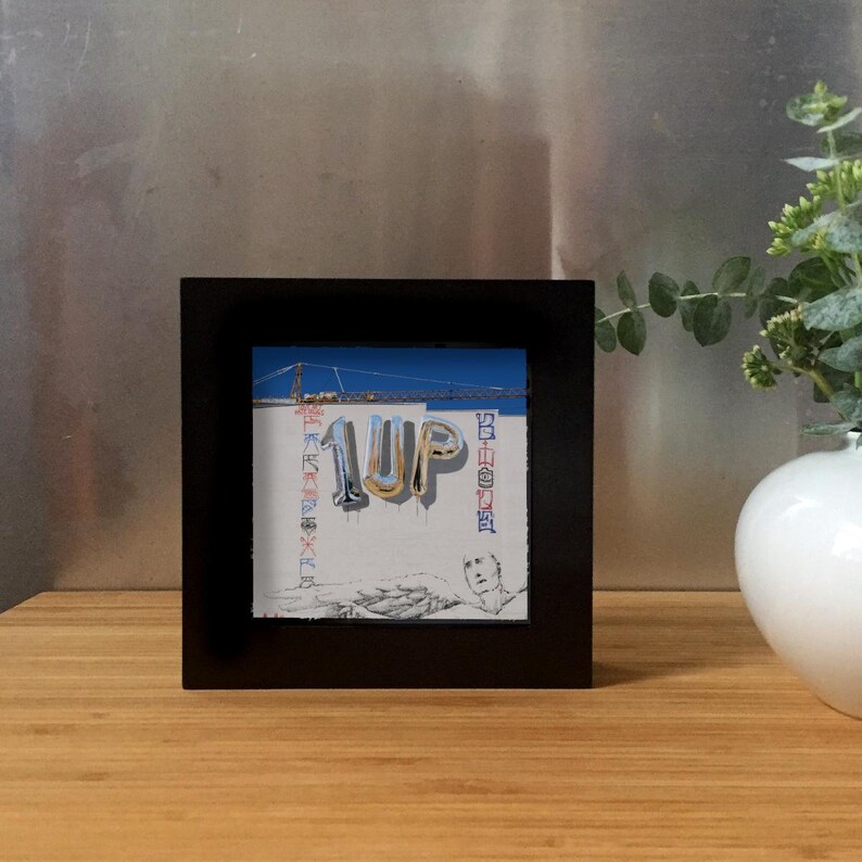 K&ouml;nnte beinhalten: Gerahmtes Kunstwerk mit dem Wort "1UP" in silbernen Ballonbuchstaben auf einer wei&szlig;en Wand mit Graffiti. Ein blauer Himmel und ein Baukran sind zu sehen. Der schwarze Rahmen umgibt das Kunstwerk.