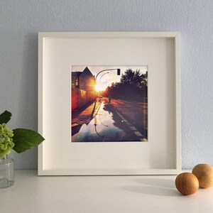 May include: A framed photograph of a city street at sunset. The image captures the sun's reflection in a puddle, with a person walking in the distance. The frame is white, and the overall aesthetic is warm and inviting.