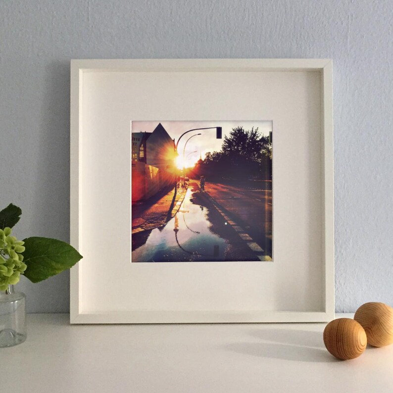 May include: A framed photograph of a city street at sunset. The image captures the sun's reflection in a puddle, with a person walking in the distance. The frame is white, and the overall aesthetic is warm and inviting.