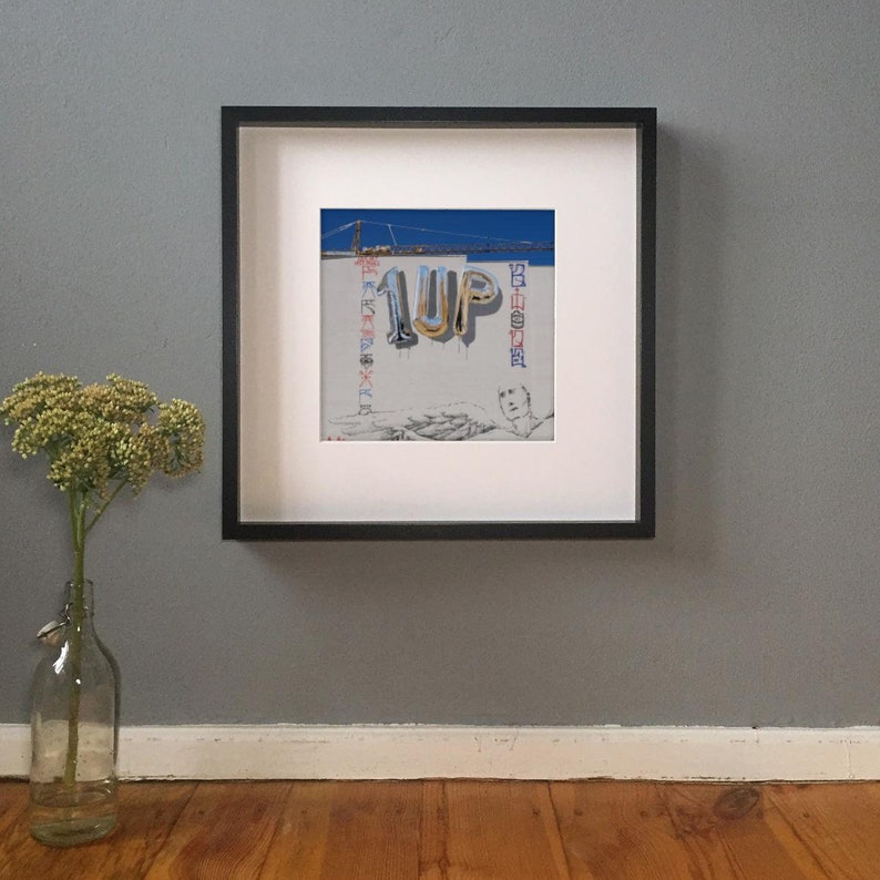 K&ouml;nnte beinhalten: Gerahmtes Kunstwerk mit einem Wandgem&auml;lde mit dem Text "1UP" in silbernen Ballonbuchstaben. Das Wandgem&auml;lde zeigt eine Br&uuml;cke und weiteren Text in verschiedenen Farben. Das Kunstwerk wird an einer grauen Wand ausgestellt.