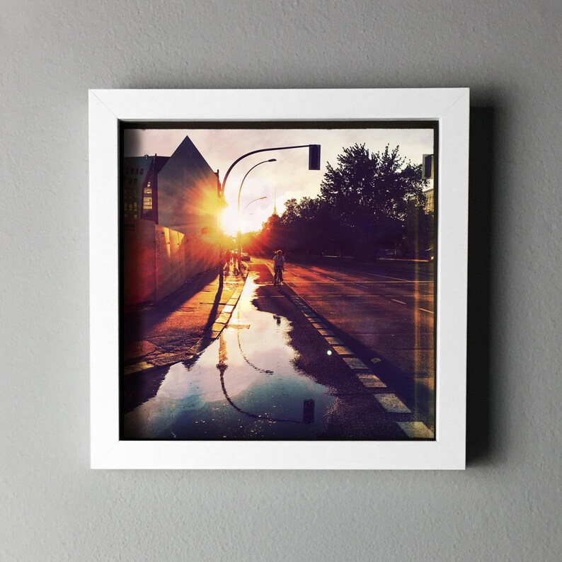 May include: Framed photograph of a city street at sunset. The sun's rays burst through the buildings, reflecting in a large puddle on the road. Silhouettes of people and streetlights are visible.