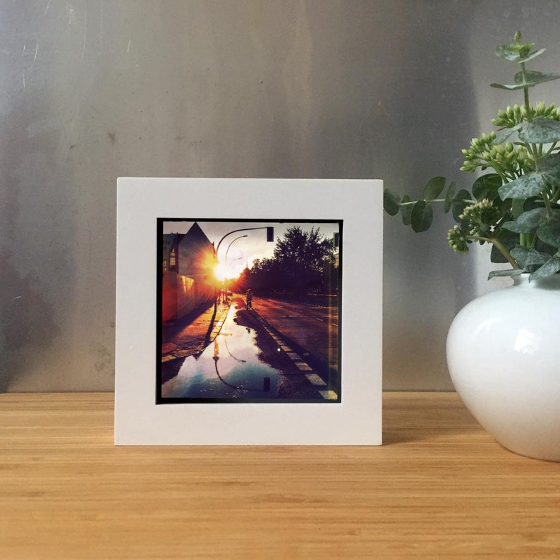 May include: A framed photograph of a street scene at sunset. The image captures a road with a person walking, reflecting the warm colors of the setting sun. The photograph is set in a white frame.