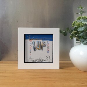 K&ouml;nnte beinhalten: Gerahmtes Kunstwerk mit wei&szlig;em Rand. Das Kunstwerk zeigt das Wort "1UP" in silbernen und goldenen Metallbuchstaben auf wei&szlig;em Hintergrund. Zus&auml;tzlicher Text und eine Zeichnung einer Person sind ebenfalls sichtbar. Der Hintergrund ist ein blauer Himmel und eine Br&uuml;cke.