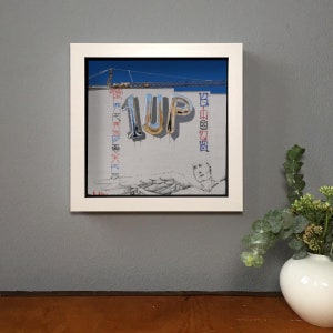 K&ouml;nnte beinhalten: Gerahmtes Kunstwerk mit dem Wort "1UP" in metallischen Ballonbuchstaben auf einer grauen Wand. Das Kunstwerk enth&auml;lt eine Zeichnung einer Person und andere Symbole. Der Rahmen ist wei&szlig; mit einem schwarzen Rand.