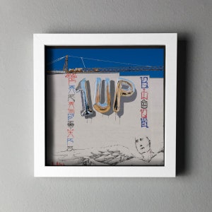 K&ouml;nnte beinhalten: Gerahmtes Kunstwerk mit dem Wort "1UP" in metallischen Ballonbuchstaben vor einer wei&szlig;en Wand. Die Wand hat Graffiti und eine Zeichnung einer Person. Ein Baukran ist vor blauem Himmel zu sehen.