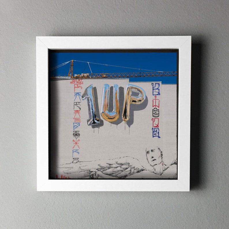K&ouml;nnte beinhalten: Gerahmtes Kunstwerk mit dem Wort "1UP" in metallischen Ballonbuchstaben vor einer wei&szlig;en Wand. Die Wand hat Graffiti und eine Zeichnung einer Person. Ein Baukran ist vor blauem Himmel zu sehen.