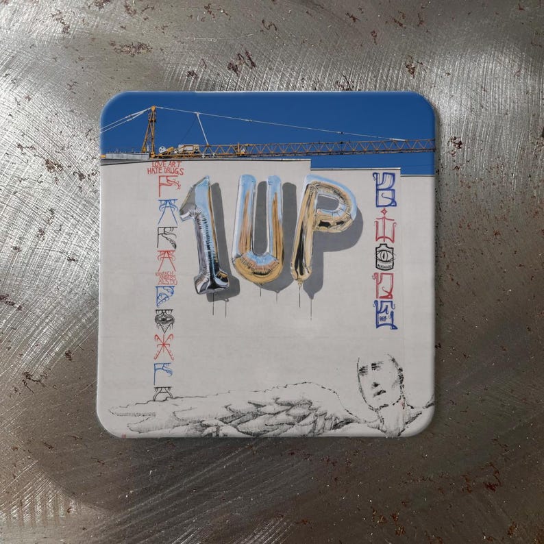 K&ouml;nnte beinhalten: Ein rechteckiger Untersetzer mit abgerundeten Ecken. Das Design zeigt den Text "1UP" in silbernen und goldenen Ballonbuchstaben. Zus&auml;tzlicher Text und Grafiken sind vorhanden, darunter ein blauer Himmel mit einem Kran und die Worte "DO NOT HATE DRUGS".