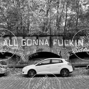 Op de afbeelding: Zwart-witfoto van een bakstenen muur met graffiti met de tekst "YOU ALL GONNA FUCKIN DIE". Drie auto's staan geparkeerd voor de muur op een geplaveide straat. De auto's zijn een zilverkleurige hatchback, een witte hatchback en een zilverkleurige sedan.