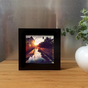 May include: A framed photograph featuring a street scene at sunset. The image captures a road reflecting the warm hues of the setting sun, with a silhouette of a person and buildings. The frame is black and square, adding a modern touch.