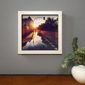 May include: A framed photograph of a city street at sunset. The image captures a cyclist on a wet road reflecting the sky's warm colors. The frame is white, and the overall scene is bathed in orange and yellow hues.