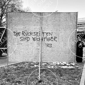 Op de afbeelding: Een zwart-witfoto van een houten muur met graffiti waarop staat "Die Ruckseiten sind wichtiger '23".