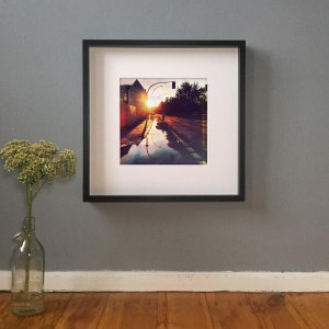 May include: Framed photograph of a city street at sunset. The image captures the sun's reflection in a puddle on the road, with a vibrant orange and yellow glow. The photograph is in a black frame with a white mat, hanging on a gray wall.