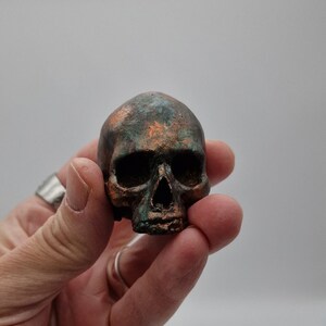 Peut inclure: Une petite sculpture de crâne peinte à la main avec une patine cuivrée et verte. Le crâne est détaillé avec des traits réalistes et a une surface texturée.