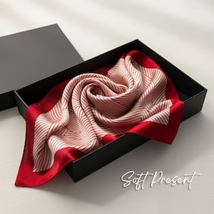 May include: A silk scarf in a black gift box. The scarf is a light pink with a geometric striped pattern and a vibrant red border. The text "Soft Present" is in the bottom right corner.