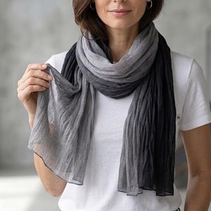 Könnte beinhalten: Ein grauer und schwarzer Ombre-Schal. Der Schal ist um Hals und Schultern drapiert. Er besteht aus einem leichten Stoff und hat eine gekreppte Textur. Ein vielseitiges Accessoire.
