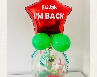 May include: A red star-shaped balloon with the text "Elijah I'M BACK" on it. The balloon is attached to a clear balloon filled with green and red confetti. There are two green balloons attached to the clear balloon.