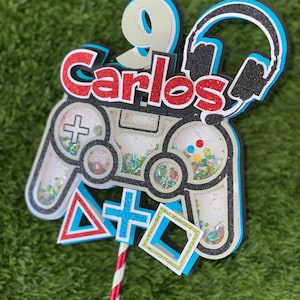 May include: A cake topper featuring a black and white video game controller with blue and green glitter accents. The controller has a red and white plus sign and a red triangle. The topper also includes a black and white headset with a blue outline, a white number nine with a blue outline, and the name "Carlos" in red letters with white outlines.