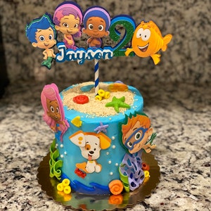 May include: A blue and white cake decorated with characters from the cartoon Bubble Guppies. The cake is topped with a topper that says "Jayson" and features a fish, a starfish, and a number 2.