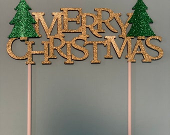 May include: Gold glitter "Merry Christmas" cake topper with two green glitter Christmas trees on either side. The topper is on two wooden sticks.