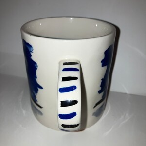 May include: White ceramic mug with a blue and black abstract design. The handle features alternating black and blue stripes. The mug has a simple, cylindrical shape and is suitable for hot or cold beverages.