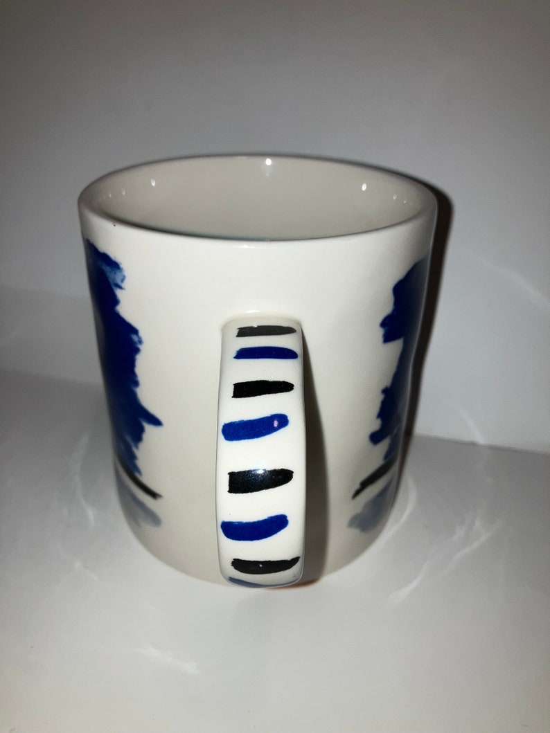May include: White ceramic mug with a blue and black abstract design. The handle features alternating black and blue stripes. The mug has a simple, cylindrical shape and is suitable for hot or cold beverages.