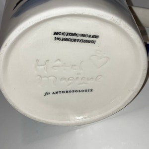May include: White ceramic mug with blue abstract design. The bottom of the mug features the embossed text "Hotel Magique" and "for ANTHROPOLOGIE". The mug is dishwasher and microwave safe.