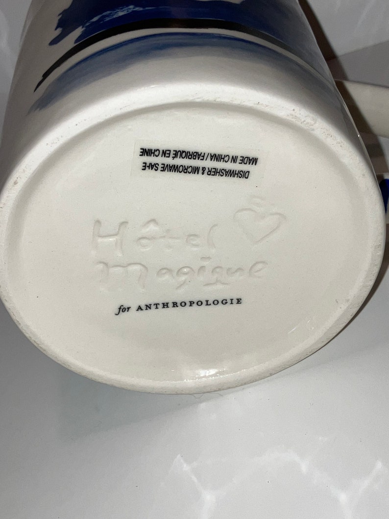 May include: White ceramic mug with blue abstract design. The bottom of the mug features the embossed text "Hotel Magique" and "for ANTHROPOLOGIE". The mug is dishwasher and microwave safe.