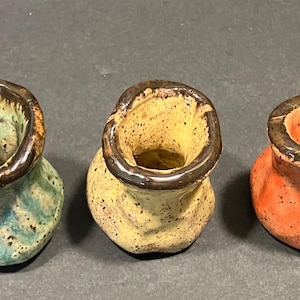 May include: Three small, handmade ceramic vases in a row. The vases are in shades of green, yellow, and orange, with a speckled texture and dark brown rims. The vases have a unique, organic shape.
