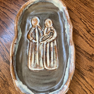 May include: A handmade ceramic dish with a raised relief depicting two figures, one holding a baby. The dish has a brown base with a lighter brown and white rim. The figures are in a light brown color.