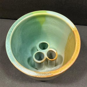 May include: A ceramic bowl with a green and brown glaze. The bowl has three holes in the bottom for holding items like pens or pencils.