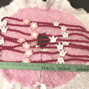May include: Several strands of red beaded garland with wooden crown and flower accents. The garland is laid out on a pink and white surface. A green ruler with the text "YOU'LL ENJOY SHOPPING" is in the foreground.