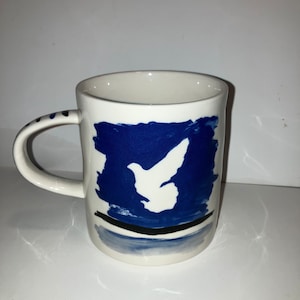 May include: White ceramic mug with a blue and white dove design. The mug features a white dove silhouette against a blue background, with a black line and blue accents at the base. The handle has blue dots.