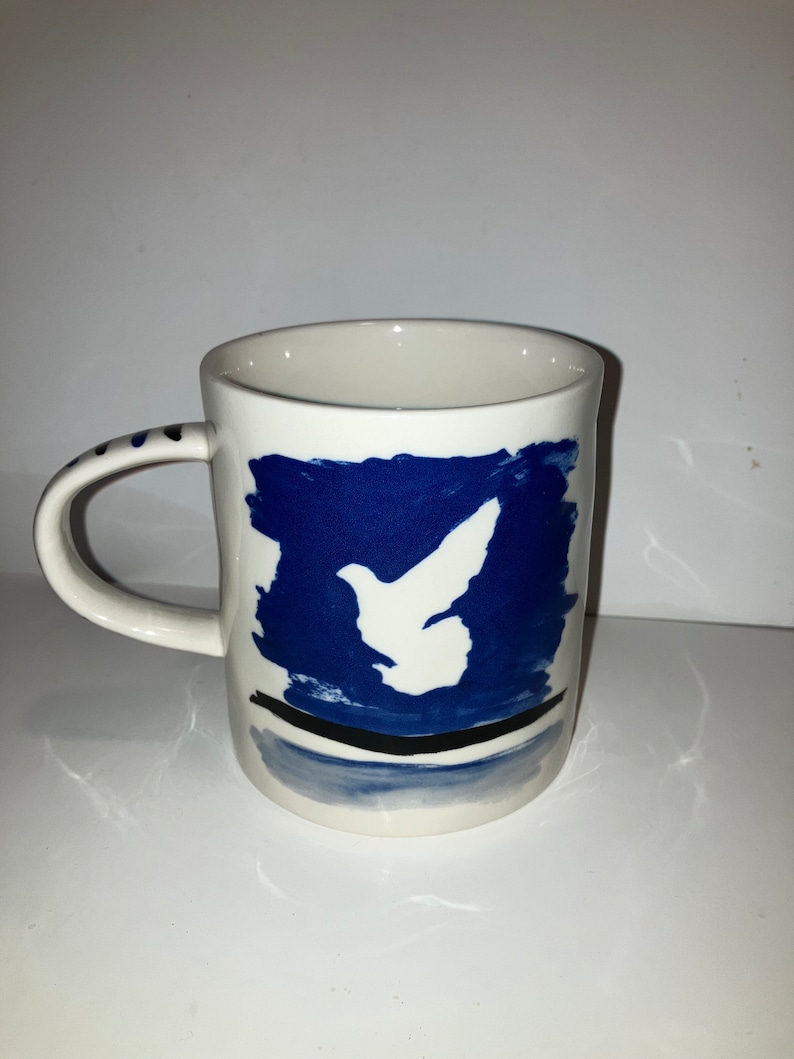 May include: White ceramic mug with a blue and white dove design. The mug features a white dove silhouette against a blue background, with a black line and blue accents at the base. The handle has blue dots.
