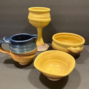 May include: A set of four handmade pottery pieces in yellow and blue. The set includes a pitcher, a goblet, and two bowls.