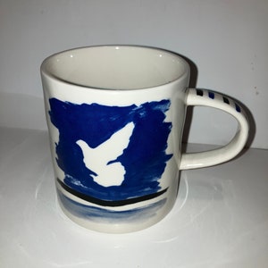 May include: White ceramic mug with a blue abstract design featuring a white dove. The handle has blue stripes. The mug is a unique piece of art.