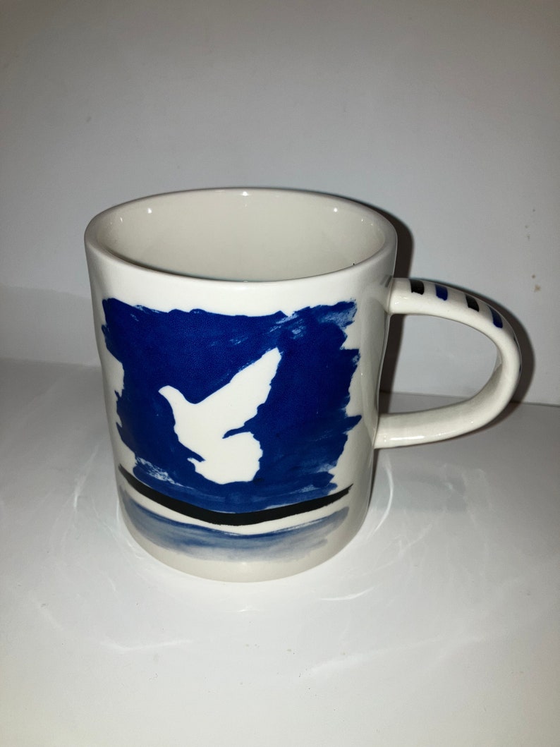 May include: White ceramic mug with a blue abstract design featuring a white dove. The handle has blue stripes. The mug is a unique piece of art.