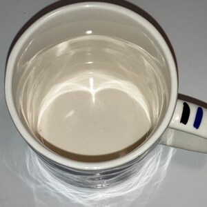May include: White ceramic mug with a rounded rim and a handle decorated with black and blue vertical stripes. The interior of the mug is a light beige color.