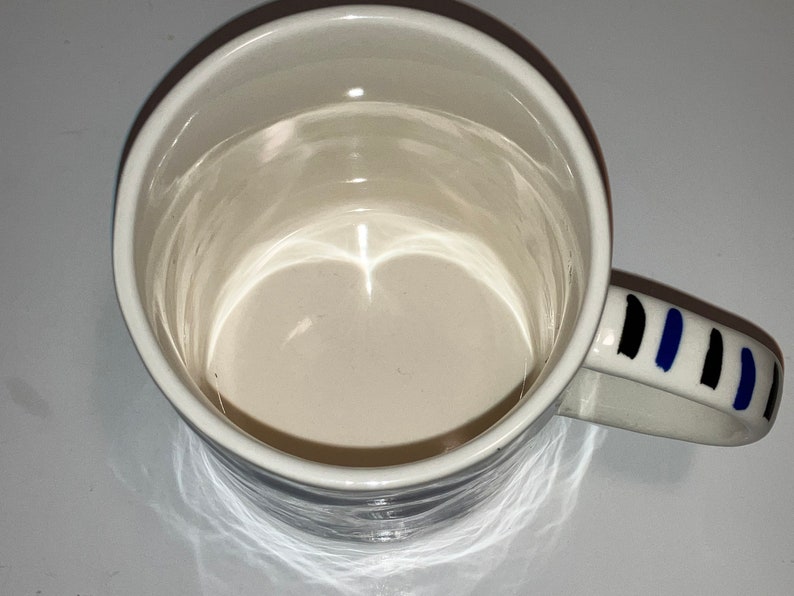 May include: White ceramic mug with a rounded rim and a handle decorated with black and blue vertical stripes. The interior of the mug is a light beige color.