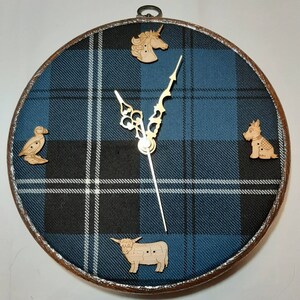 May include: A round clock with a blue and black plaid design. The clock has a silver frame and wooden hands. The clock face is decorated with four wooden cutouts of a unicorn, a puffin, a highland cow, and a terrier.