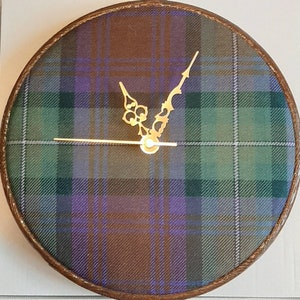 May include: A round clock with a brown wooden frame and a plaid fabric face. The clock hands are gold and have a decorative design.